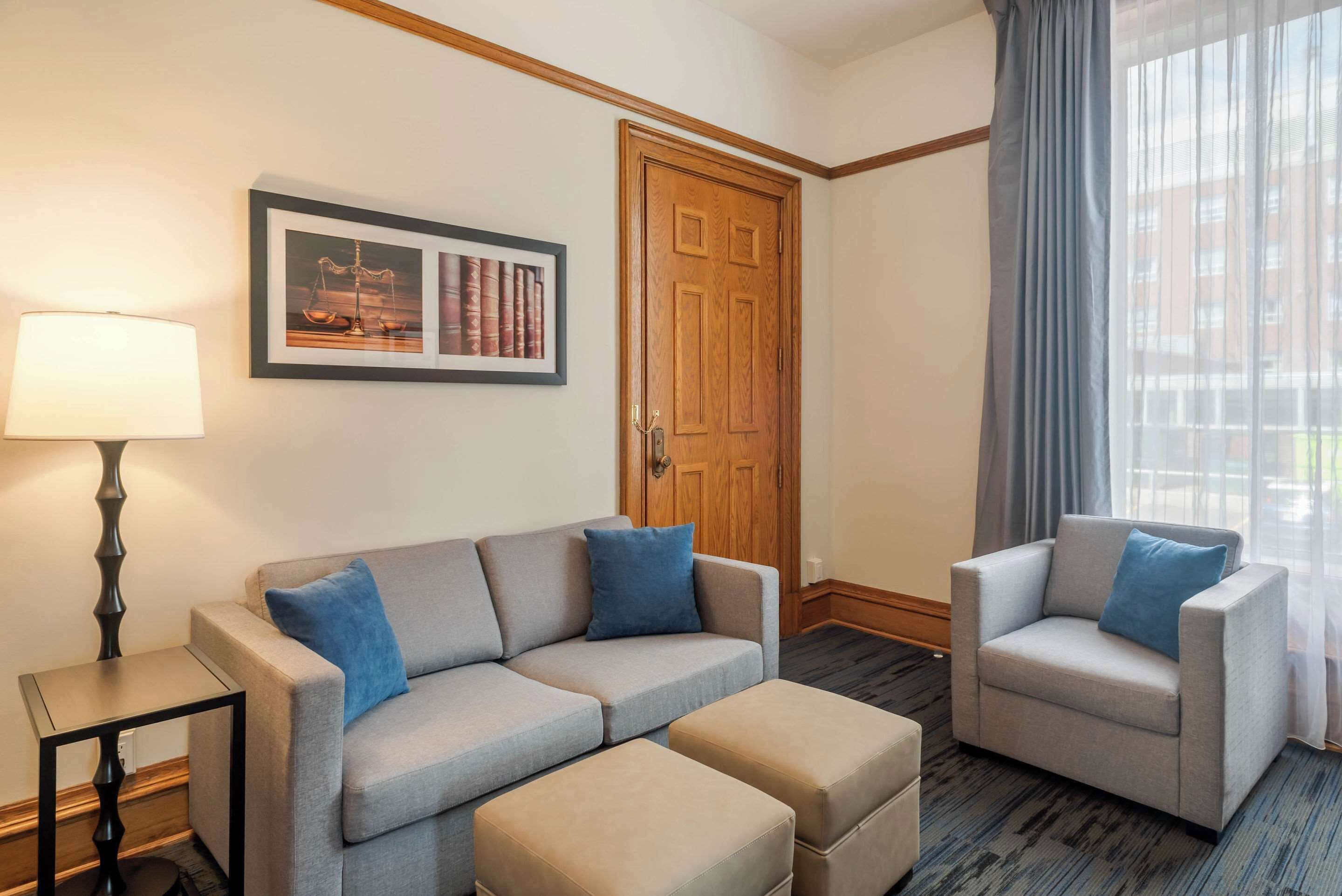 Camera d'albergo con un divano grigio, una poltrona abbinata, un tavolino e una lampada. Si nota un quadro incorniciato appeso al muro e una porta in legno.