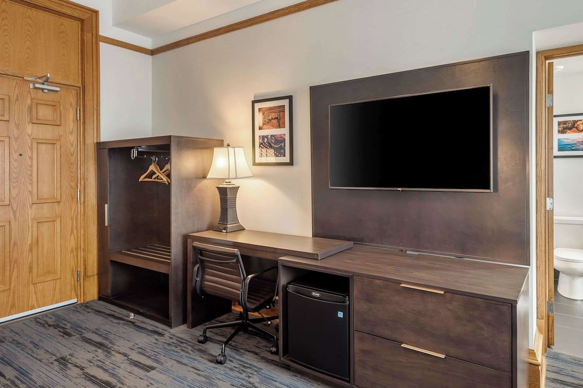 A hotel room with a wooden wardrobe, a desk with a lamp, a television, and a small refrigerator.