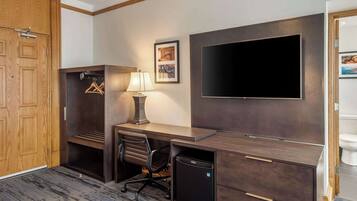 A hotel room with a wooden wardrobe, a desk with a lamp, a television, and a small refrigerator.