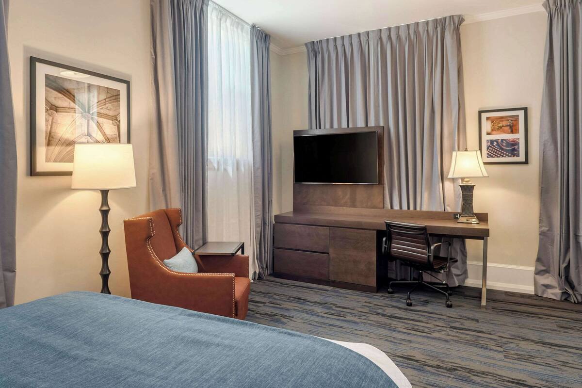 A hotel room with a bed, a desk with a chair, a TV, and a lamp.