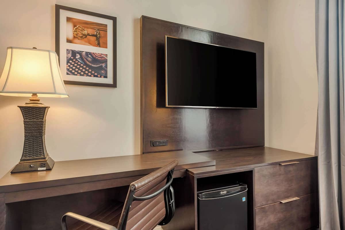 A hotel room with a flat-screen TV mounted on the wall, a desk with a lamp, a chair, and a small black appliance, possibly a mini-fridge or heater.
