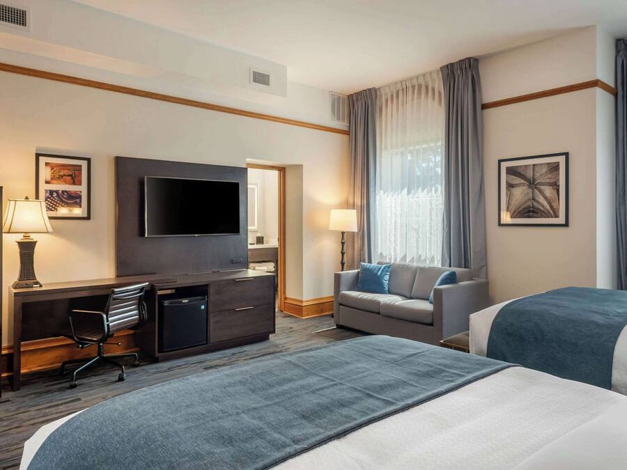 Habitación de hotel con una cama grande, un escritorio con silla, televisión, sofá y ventana con cortinas.