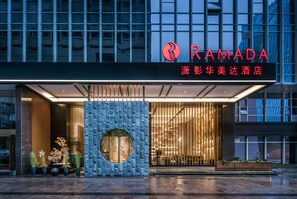 L'entrée d'un hôtel moderne, avec un logo bien visible, une façade en pierre et un intérieur bien éclairé, que l'on peut apercevoir à travers de grandes fenêtres.