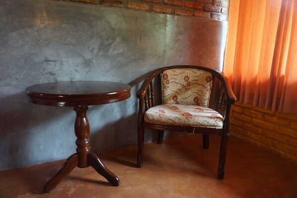 Una silla de madera con tapicería floral y una mesa redonda de madera con base de trípode, ubicada frente a una pared de ladrillo y una ventana con cortinas.