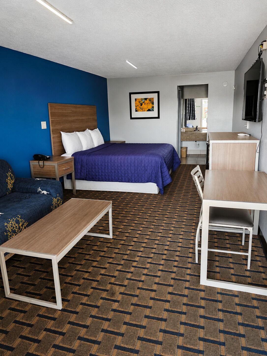 A hotel room with a bed, a desk, a chair, a sofa, a TV, and a bathroom visible in the background.