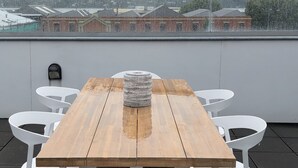 A wooden table with white chairs on a rooftop terrace with a city view. - Bond Street Apartment Luxury 3-bedroom (Dunedin)