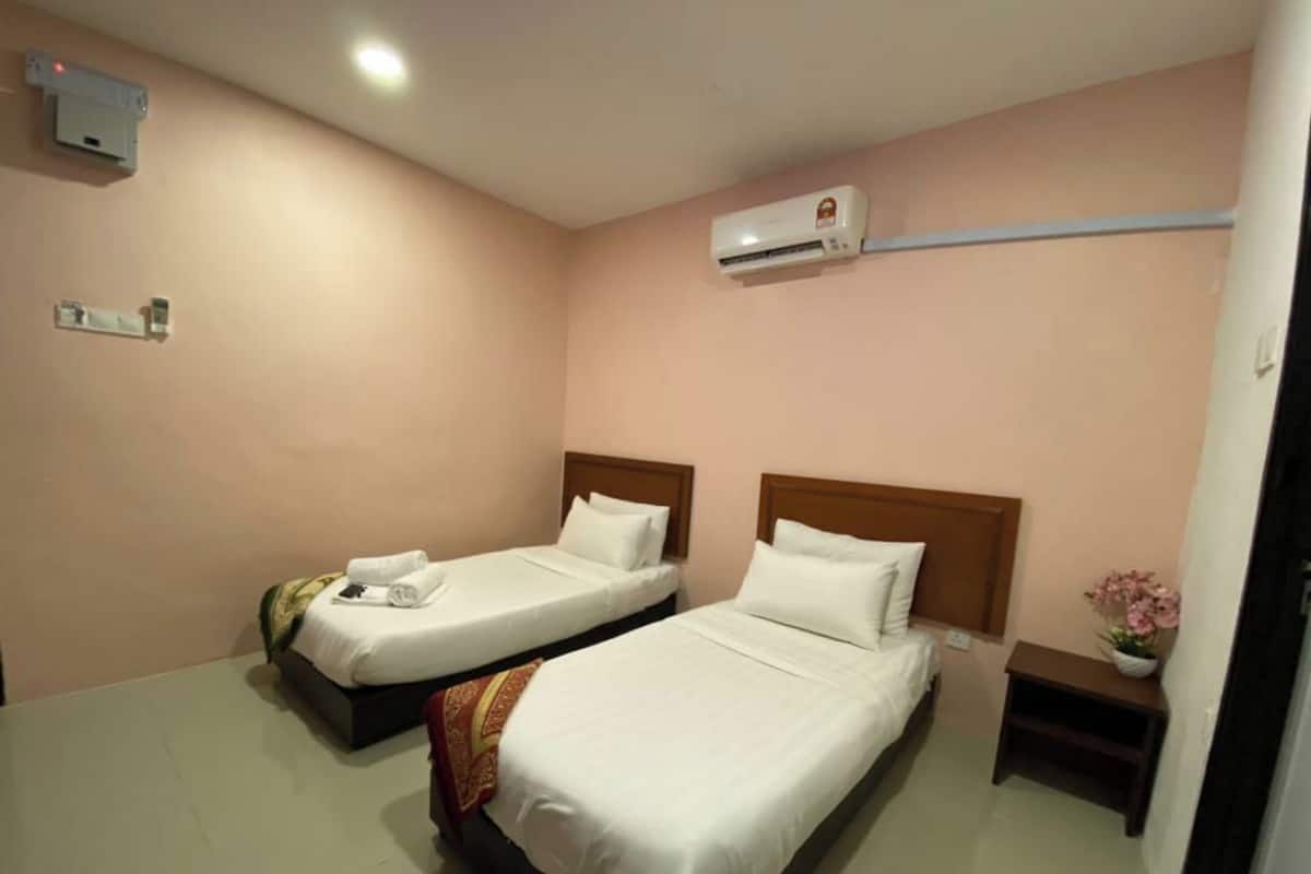 Habitación de hotel con dos camas, un equipo de aire acondicionado, una mesita de noche con un jarrón de flores y una lámpara fijada a la pared.
