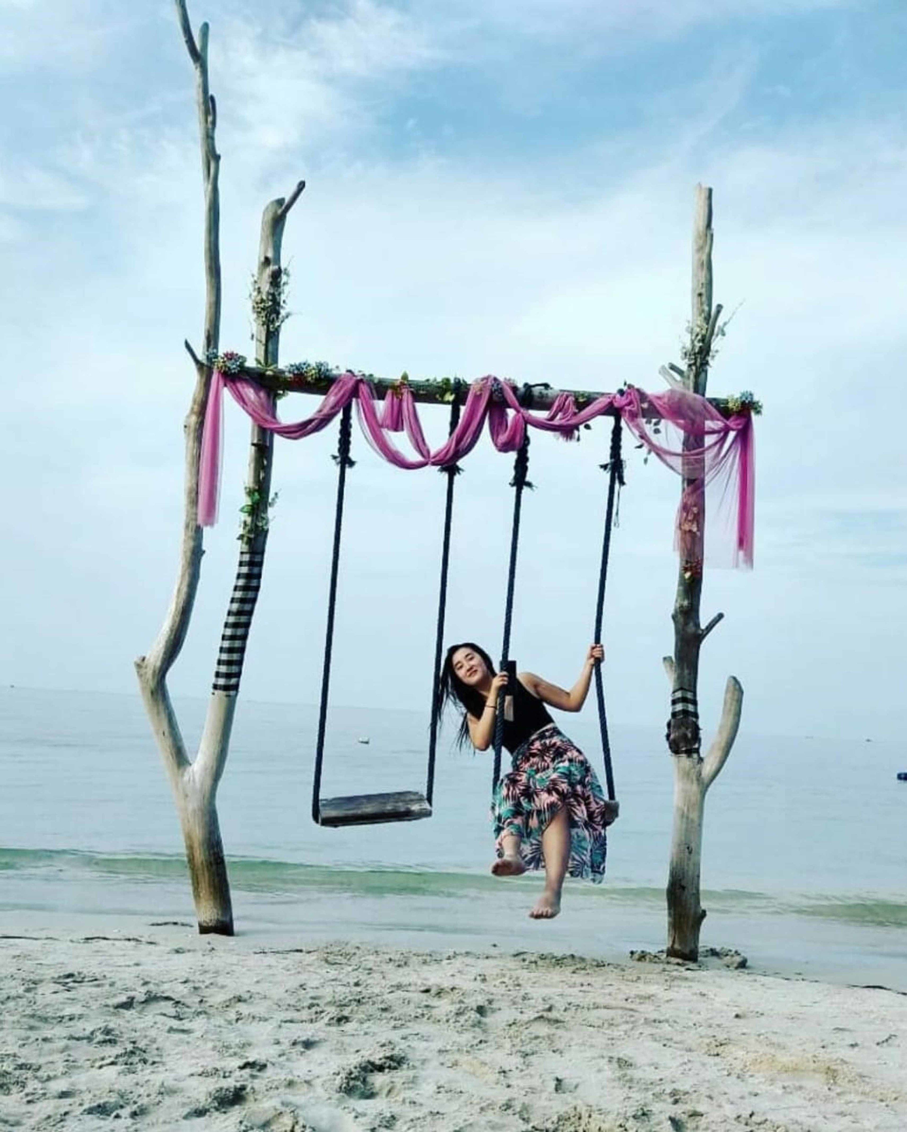 Columpios en la playa, con una persona balanceándose, rodeados de árboles y un cielo despejado.