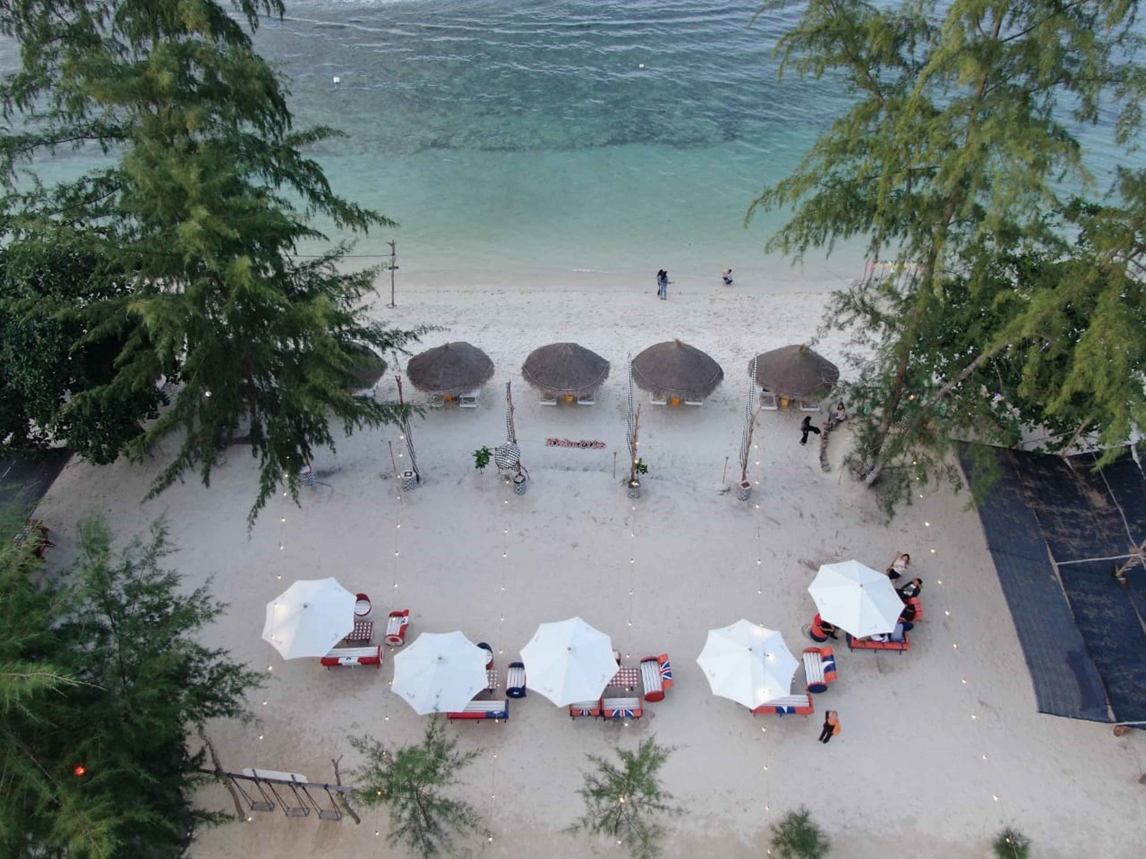 Une plage avec des cabanes aux toits de chaume et des parasols, dont certains sont ornés de drapeaux, et quelques personnes qui se promènent.