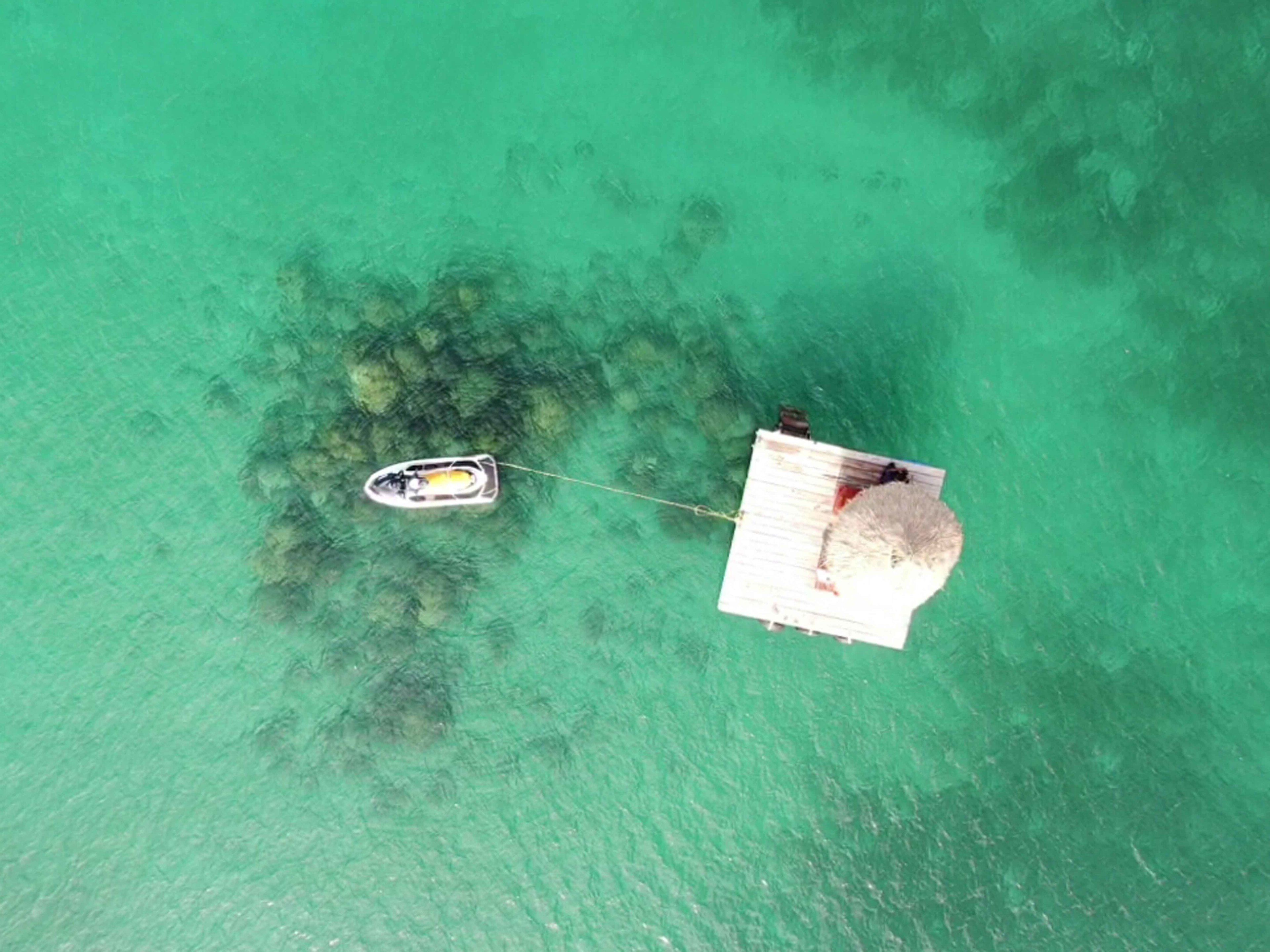 Ein Luftbild zeigt ein Boot, das an einem Steg angebunden ist, mit klarem türkisblauem Wasser.