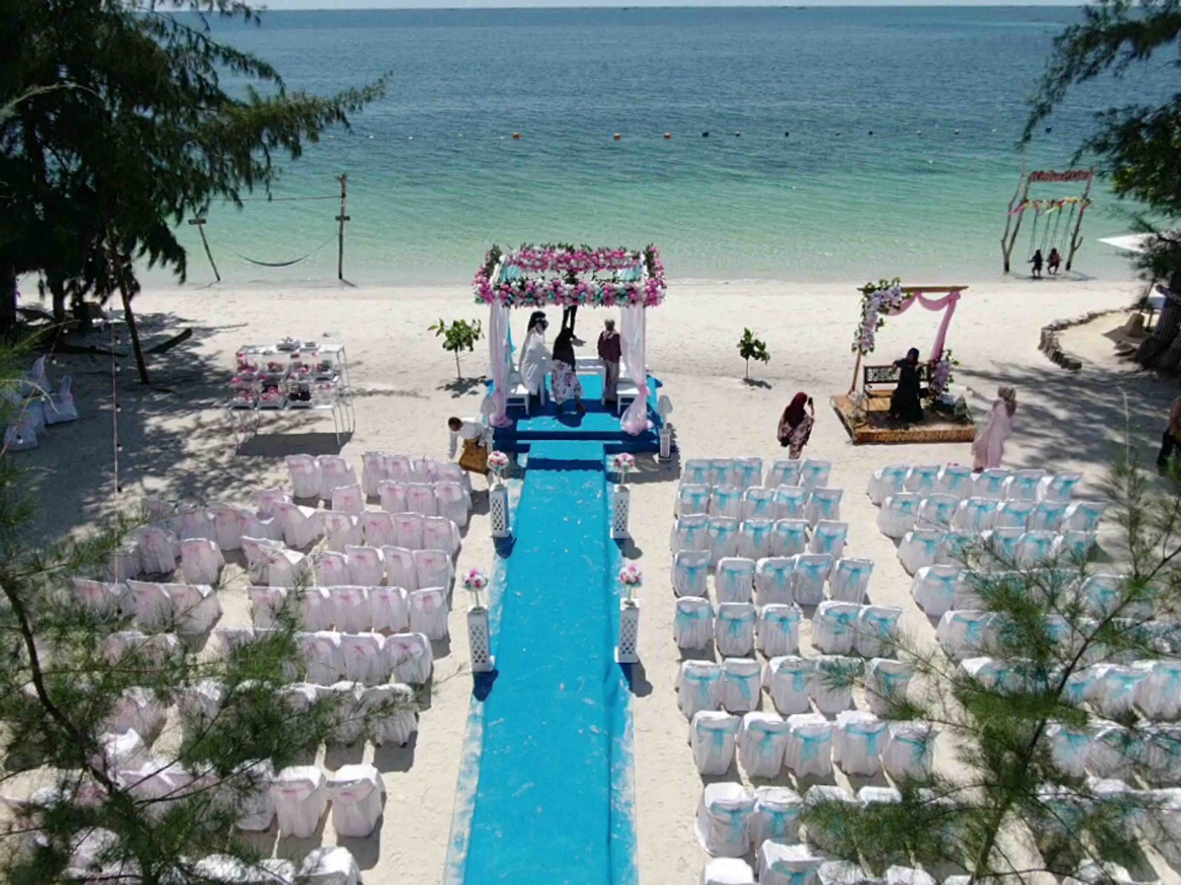 A beach wedding ceremony with a blue aisle, white chairs, and a floral arch.