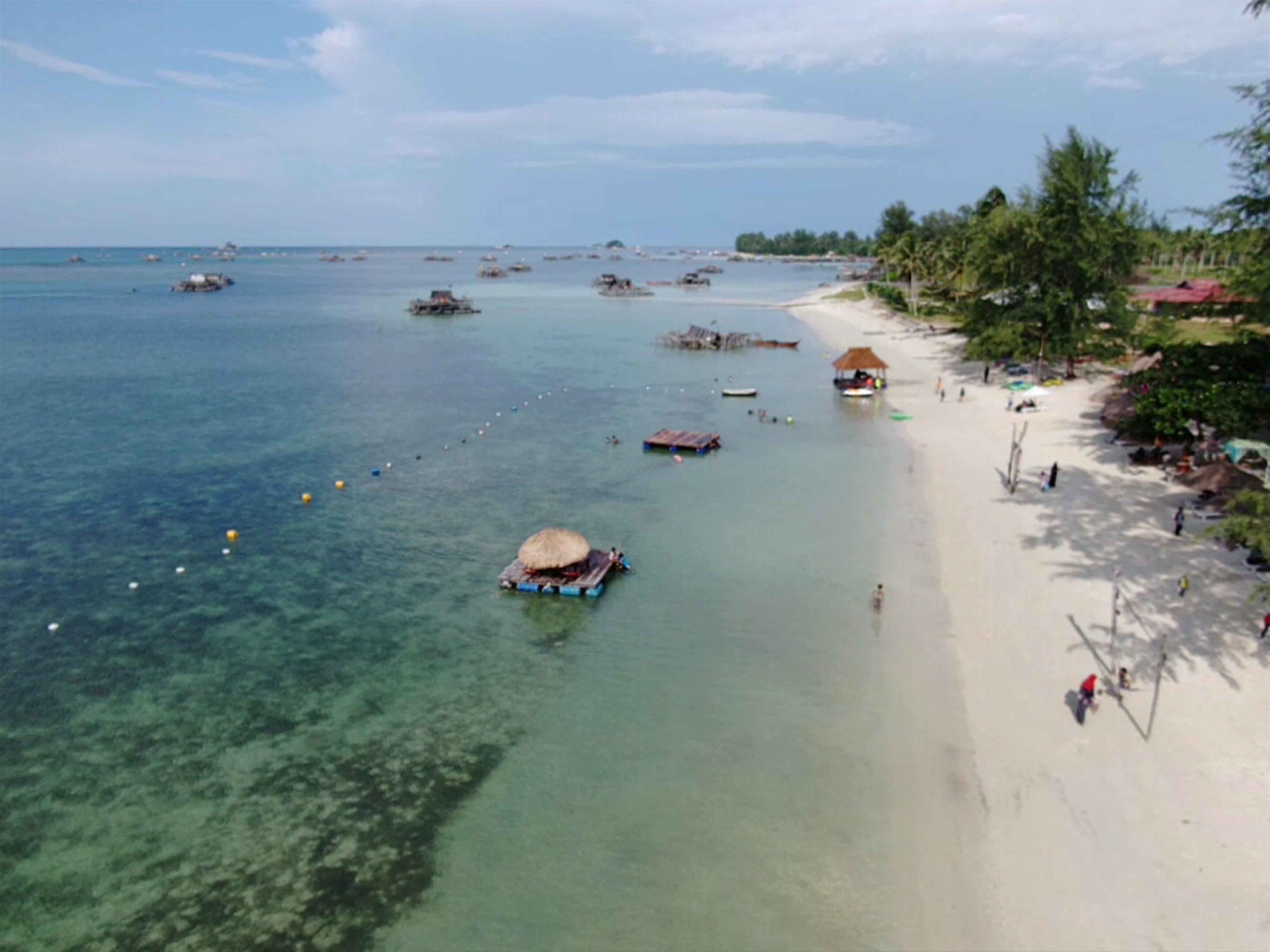A beach with clear shallow waters, boats, and a few huts.