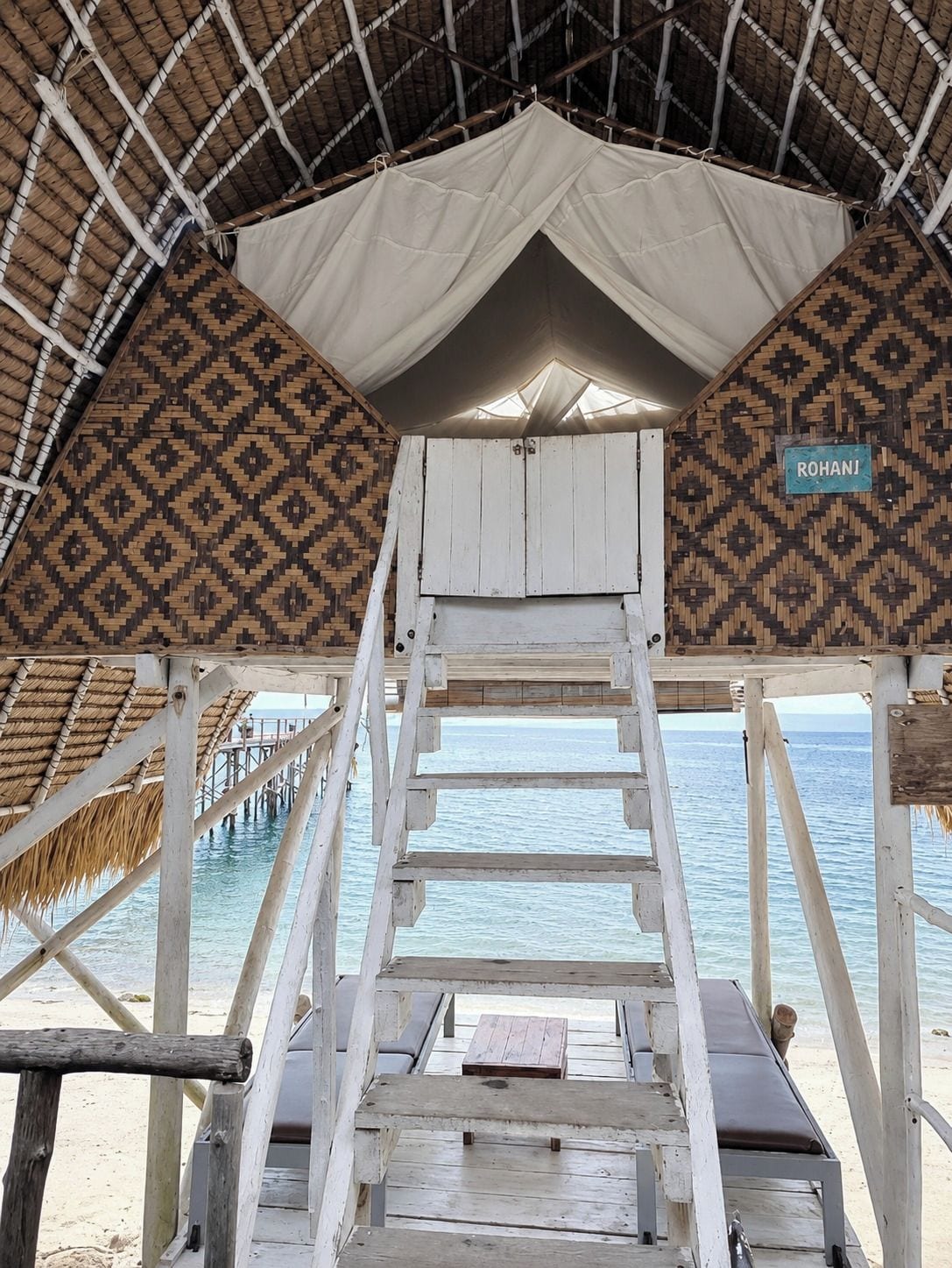 Una terraza de madera con techo de paja, un cartel que dice 'ROHANI' y vista al mar.
