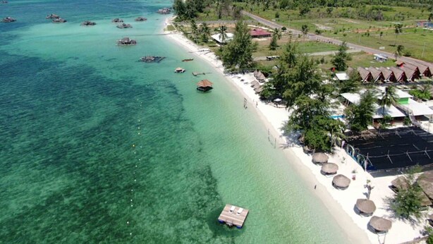 Luchtbeeld van een strandresort met helder turquoise water, een zandstrand en verschillende bungalows boven het water.