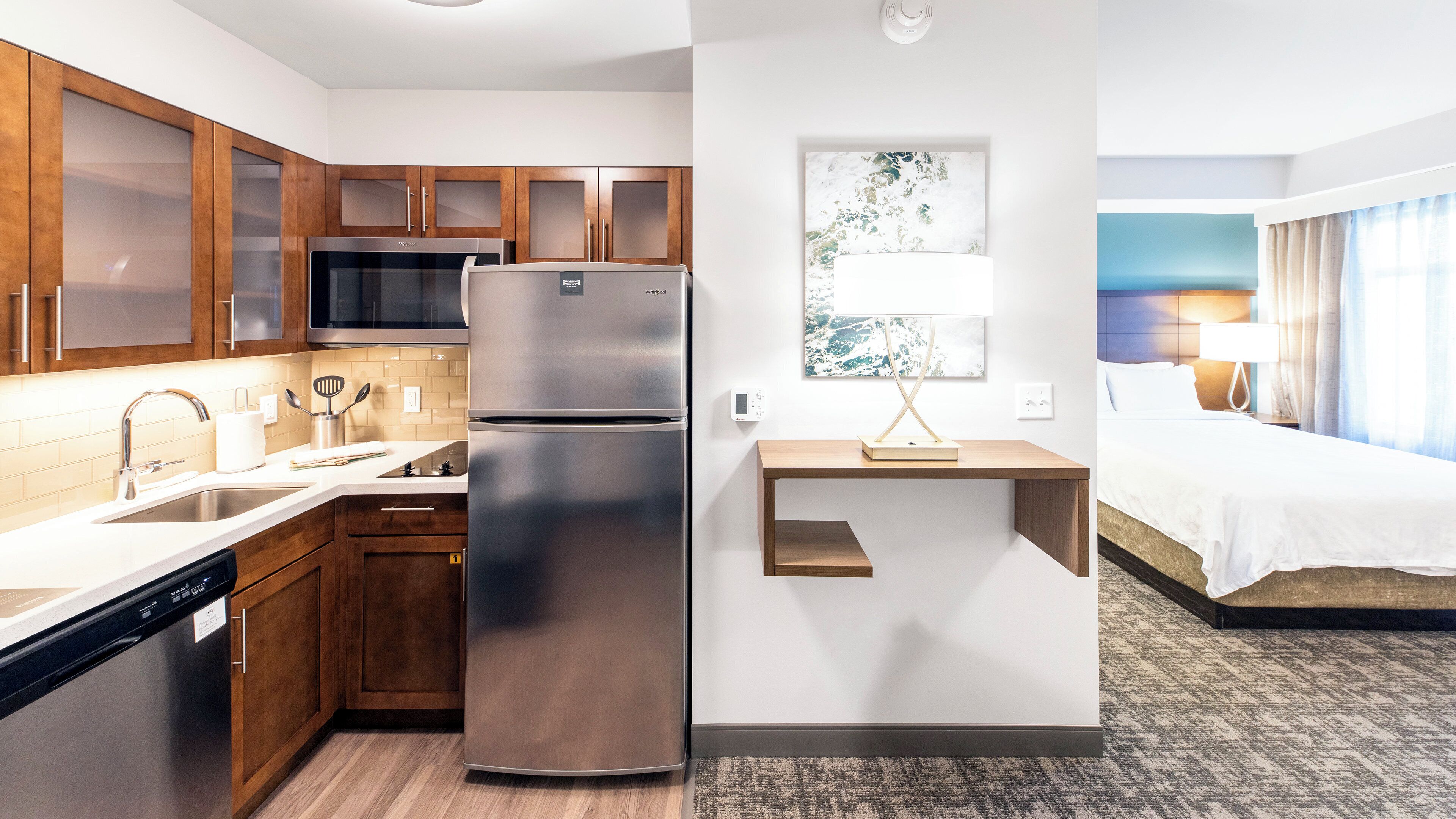 A modern hotel room with a kitchenette, a bed, a bedside table, and a wall-mounted lamp.
