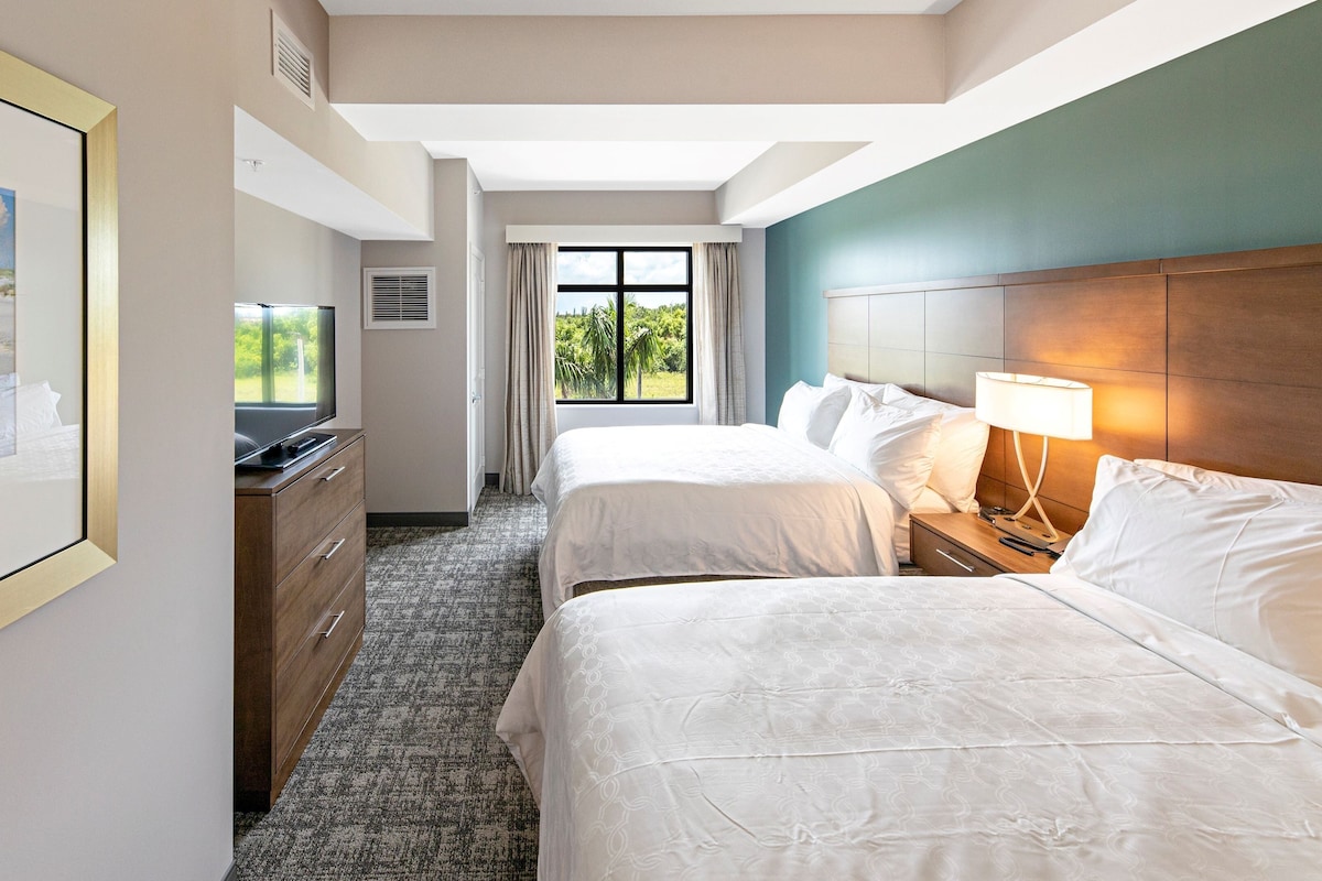 A hotel room with two beds, a desk with a TV, a window with a view of greenery, and a framed picture on the wall.
