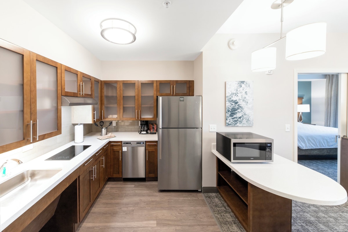 A modern kitchen with wooden cabinets, stainless steel appliances, a microwave, and a view of a bedroom.