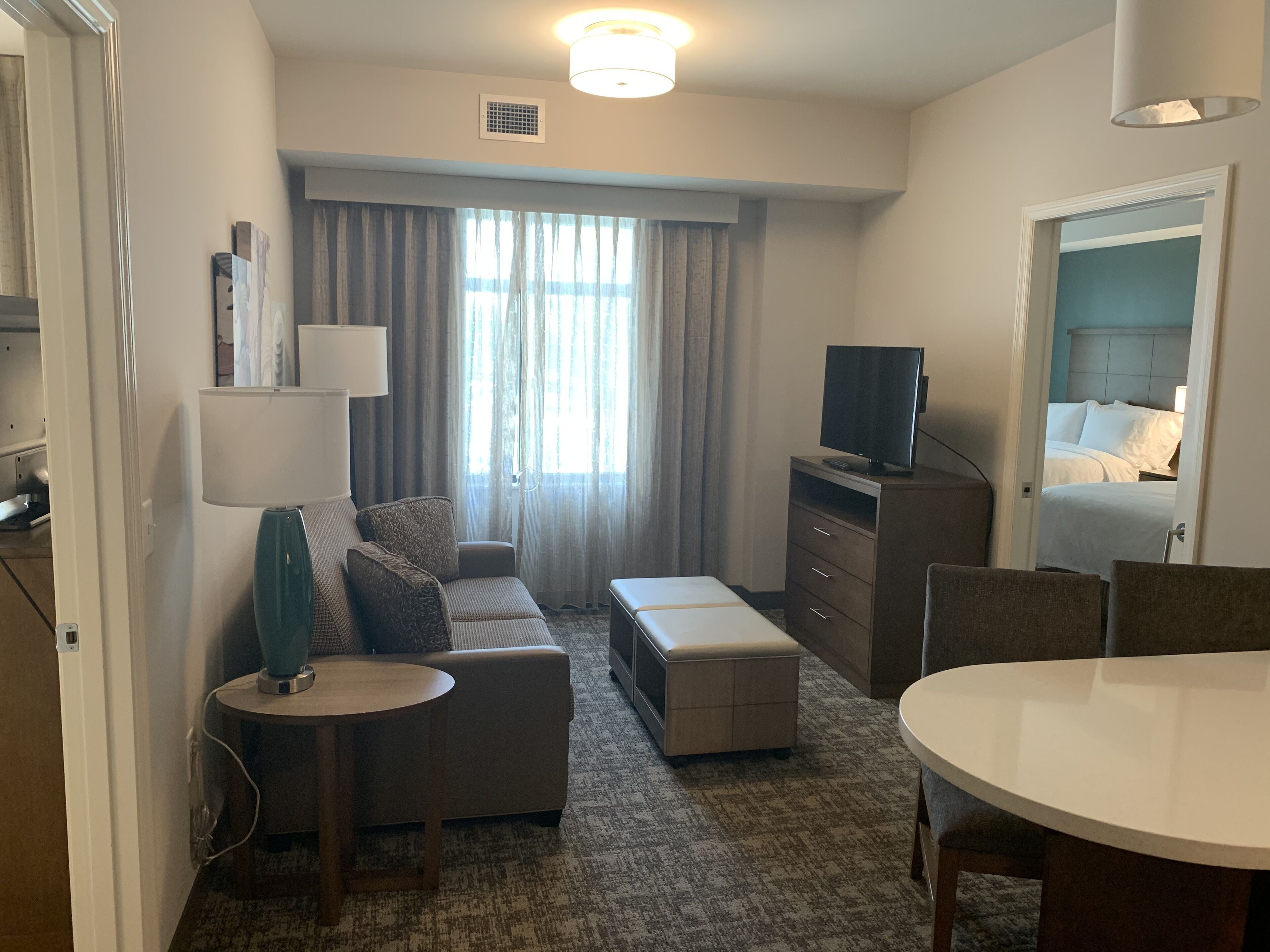 A hotel room with a sofa, a small table, a TV on a dresser, a bed, and a window with curtains.