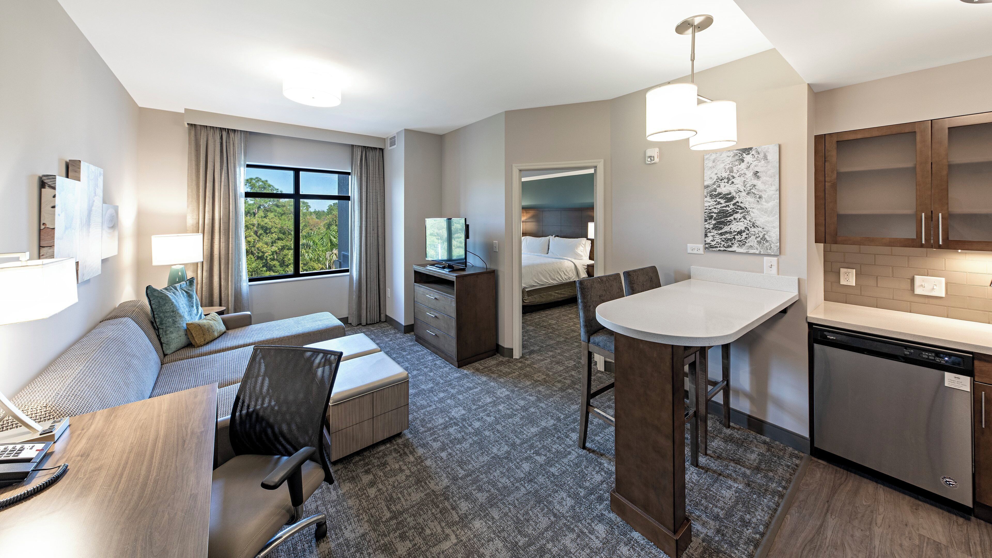 A hotel room with a bed, a desk, a chair, a kitchenette, and a view of the outdoors.