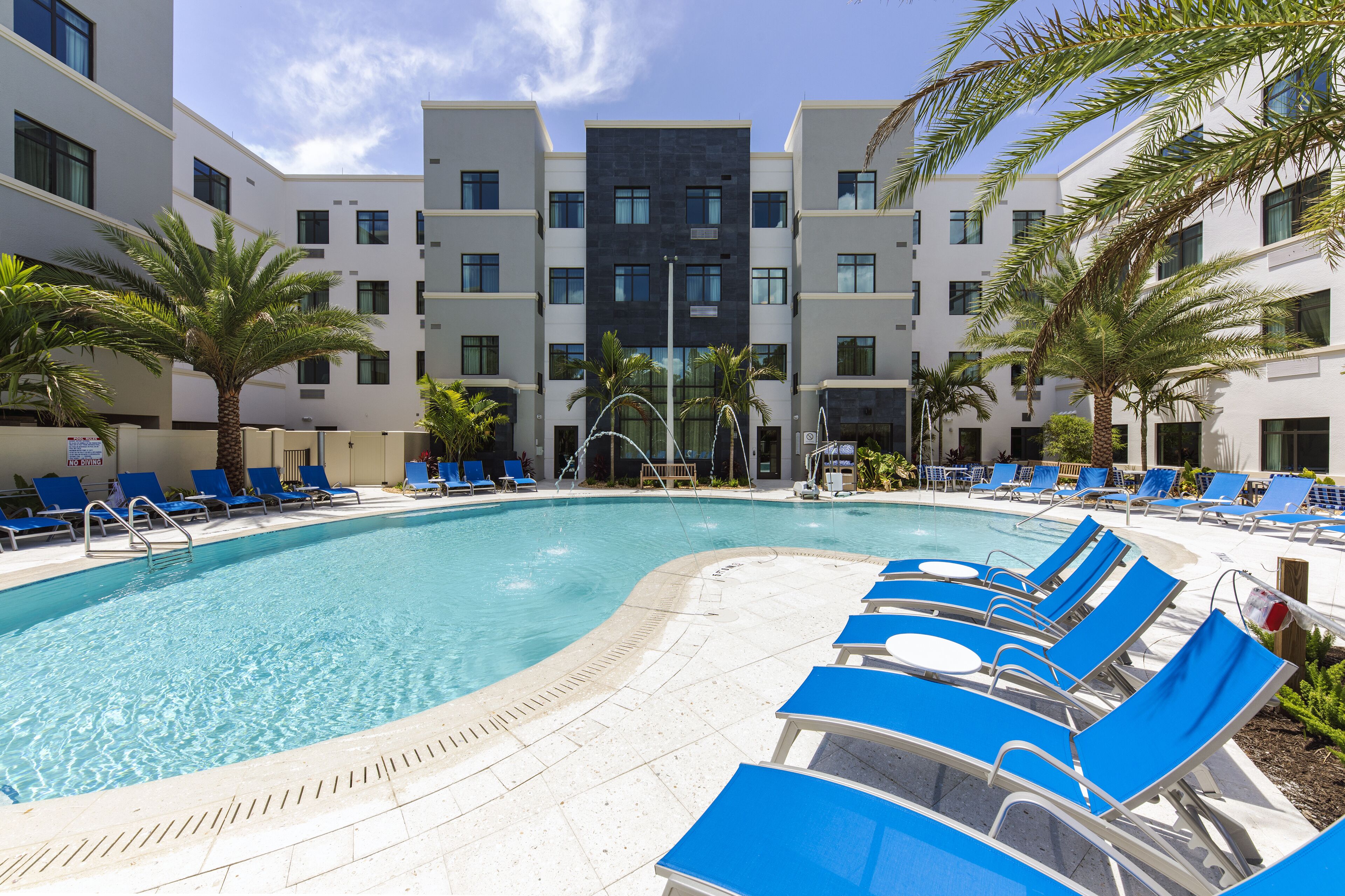 A swimming pool with blue lounge chairs and palm trees, surrounded by modern buildings.