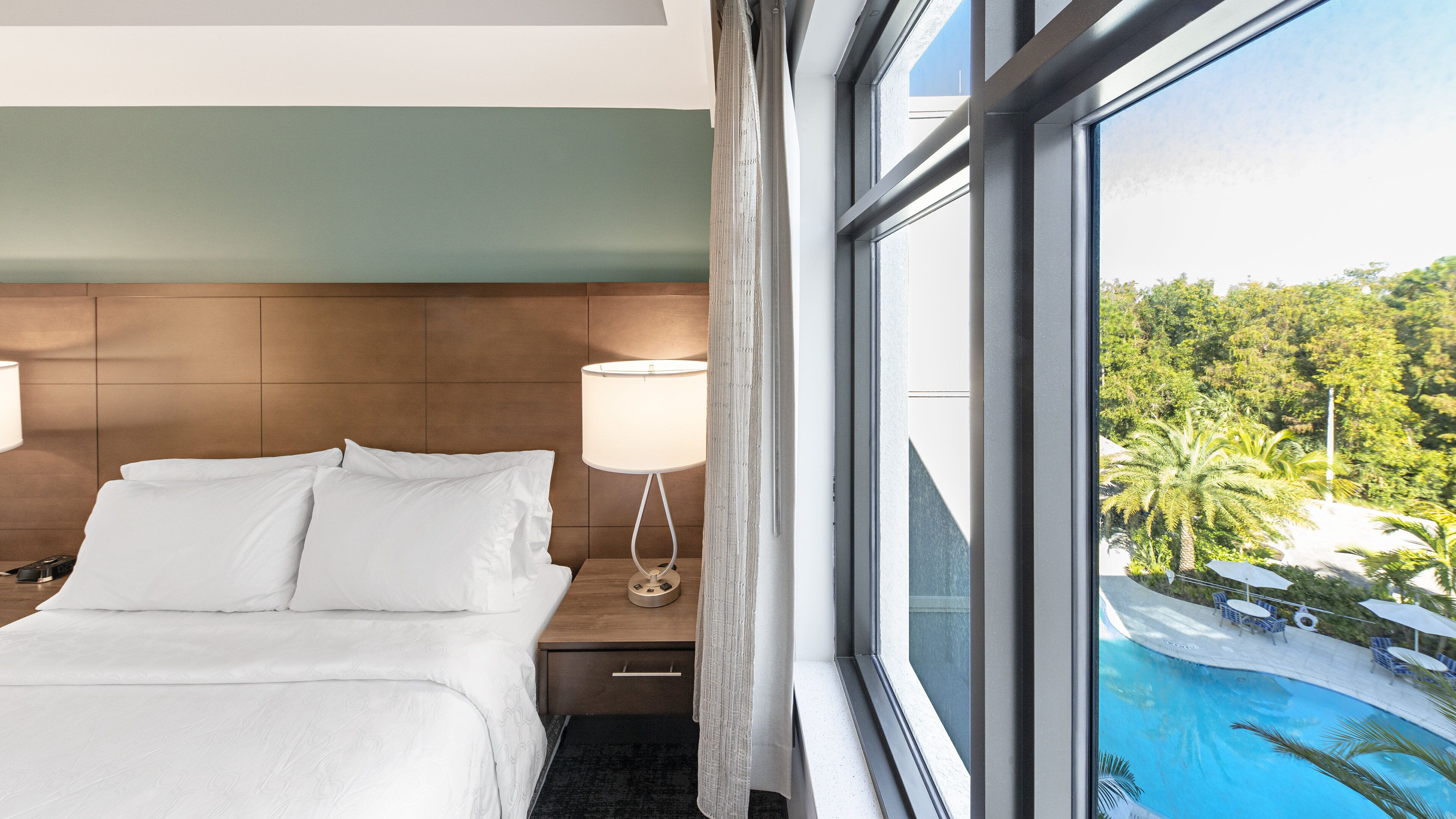 Une chambre d’hôtel avec un lit, une table de chevet, une lampe et une fenêtre donnant sur une piscine et un espace verdoyant.