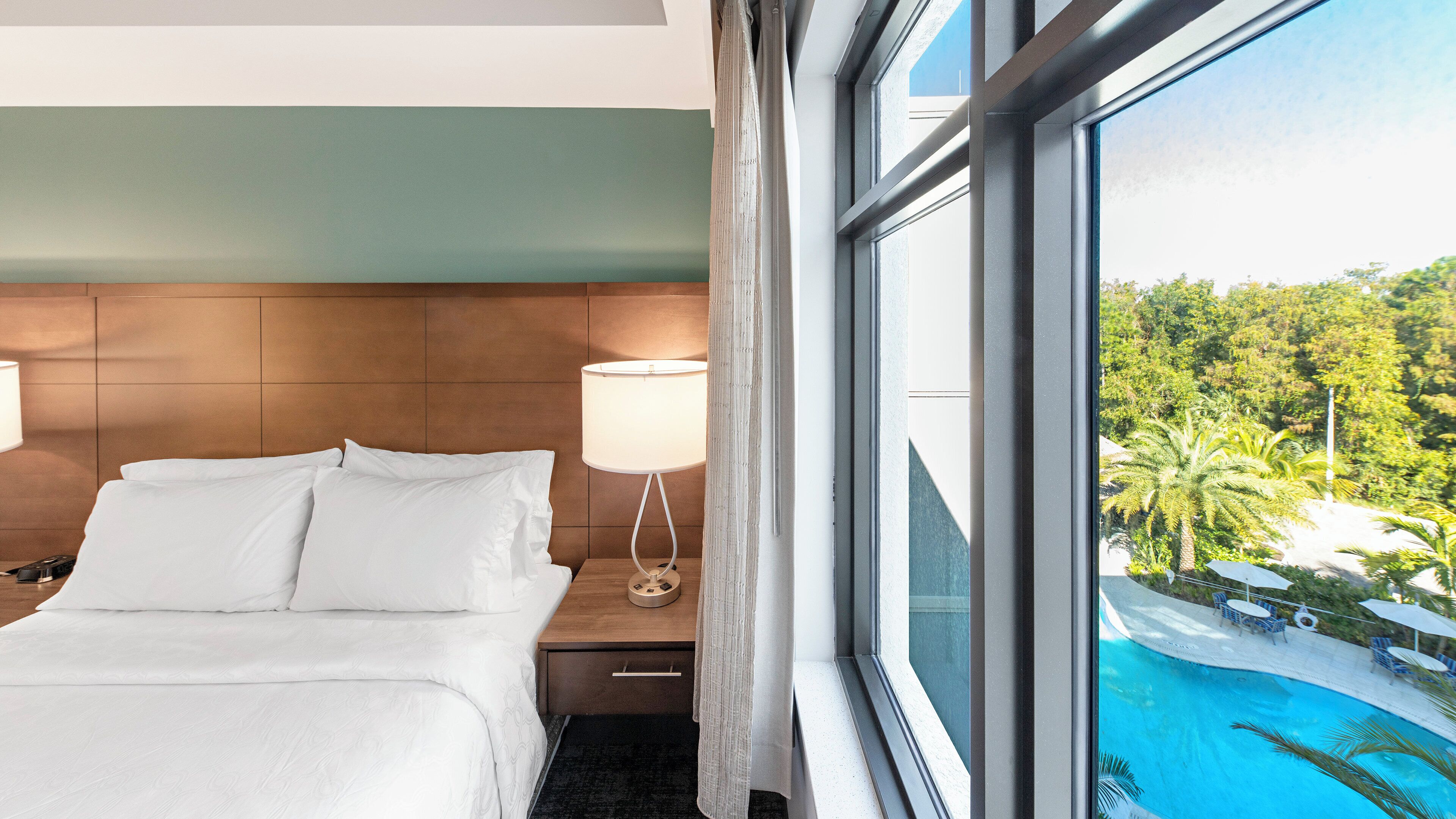 A hotel room with a bed, a nightstand, a lamp, and a view of a swimming pool and greenery.