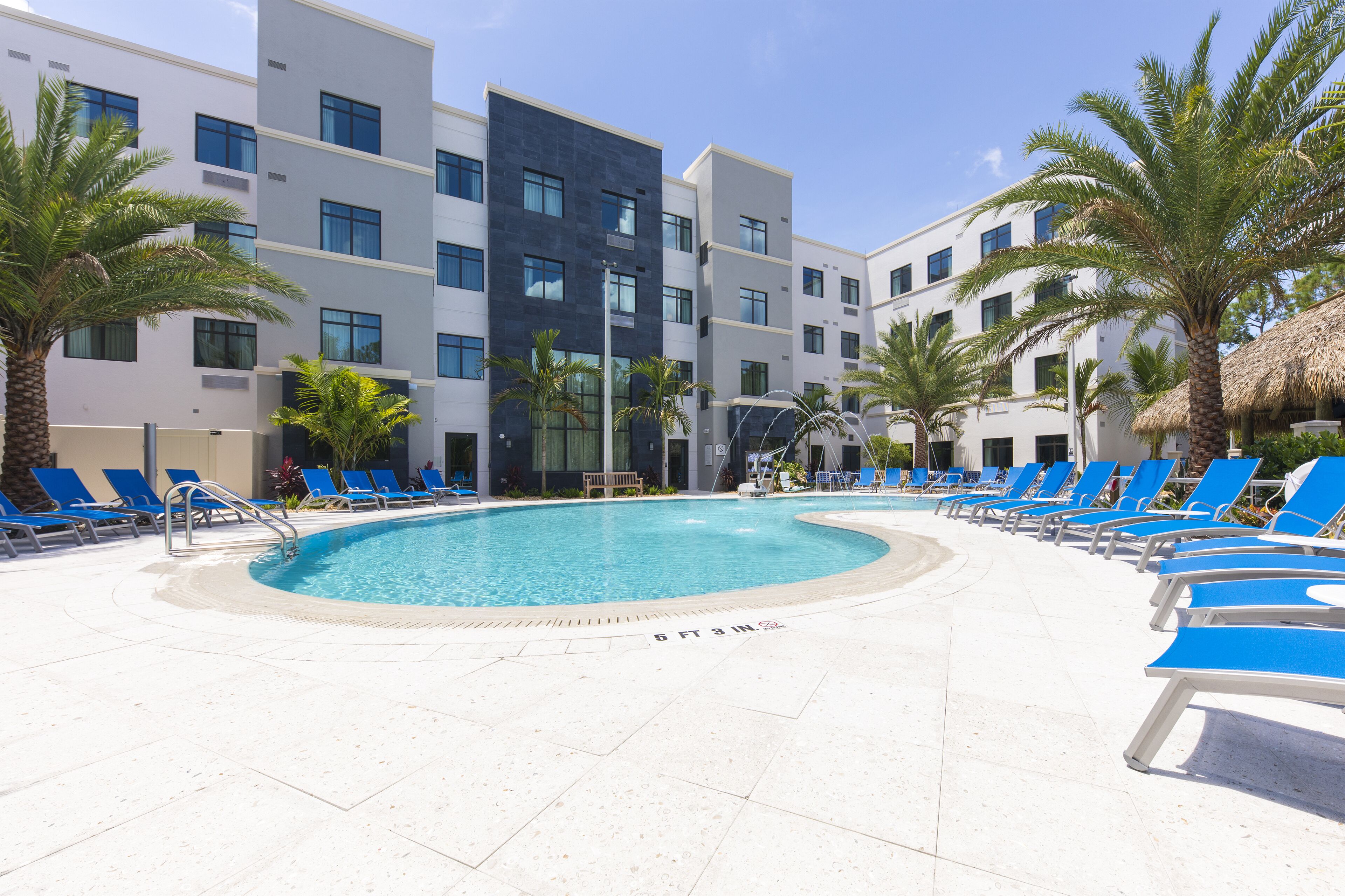 A swimming pool surrounded by blue lounge chairs, palm trees, and modern buildings with multiple floors.