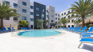 A swimming pool surrounded by blue lounge chairs, palm trees, and modern buildings with multiple floors.