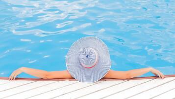 Una persona relajándose junto a una piscina con un sombrero a rayas, una cubierta de madera y agua cristalina azul.