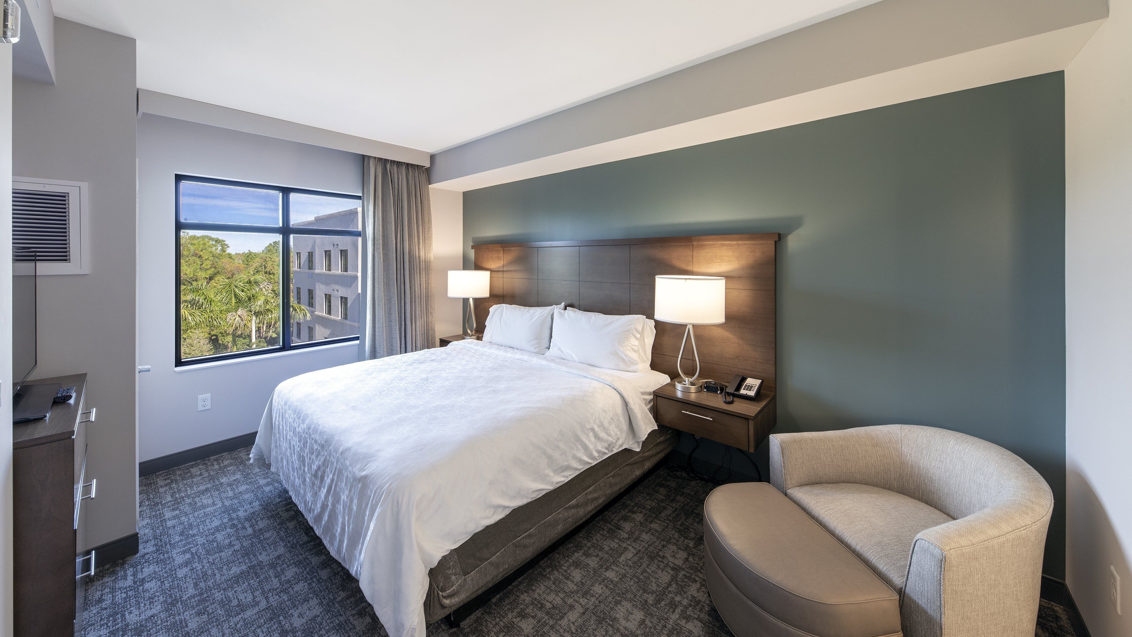 Ein Hotelzimmer mit einem großen Bett, zwei Nachttischlampen, einem Sessel, einem Schreibtisch mit einem Laptop und einem Fenster mit Blick auf Gebäude und Bäume.