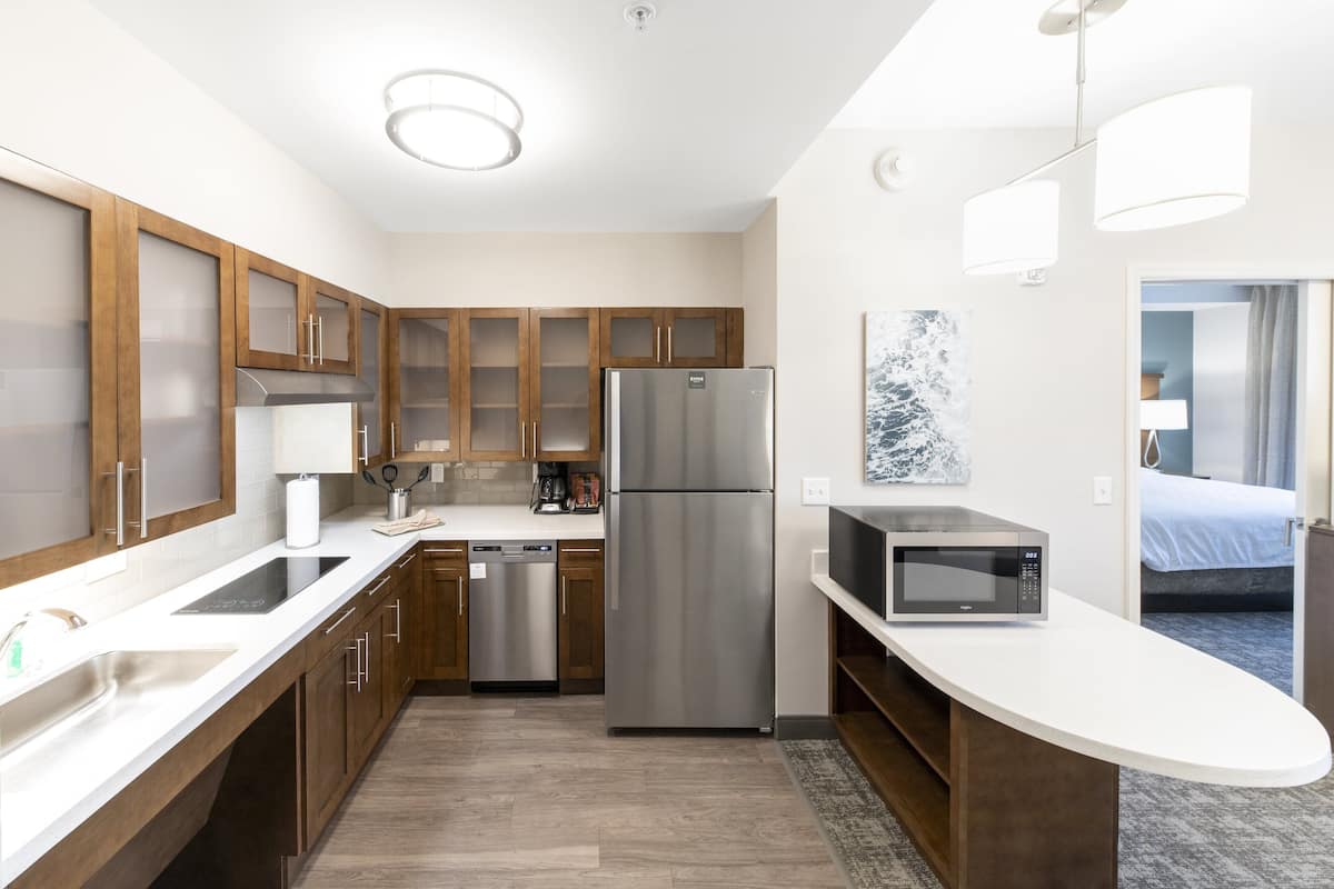 A modern kitchen with wooden cabinets, stainless steel appliances, a microwave, and a view of a bedroom.