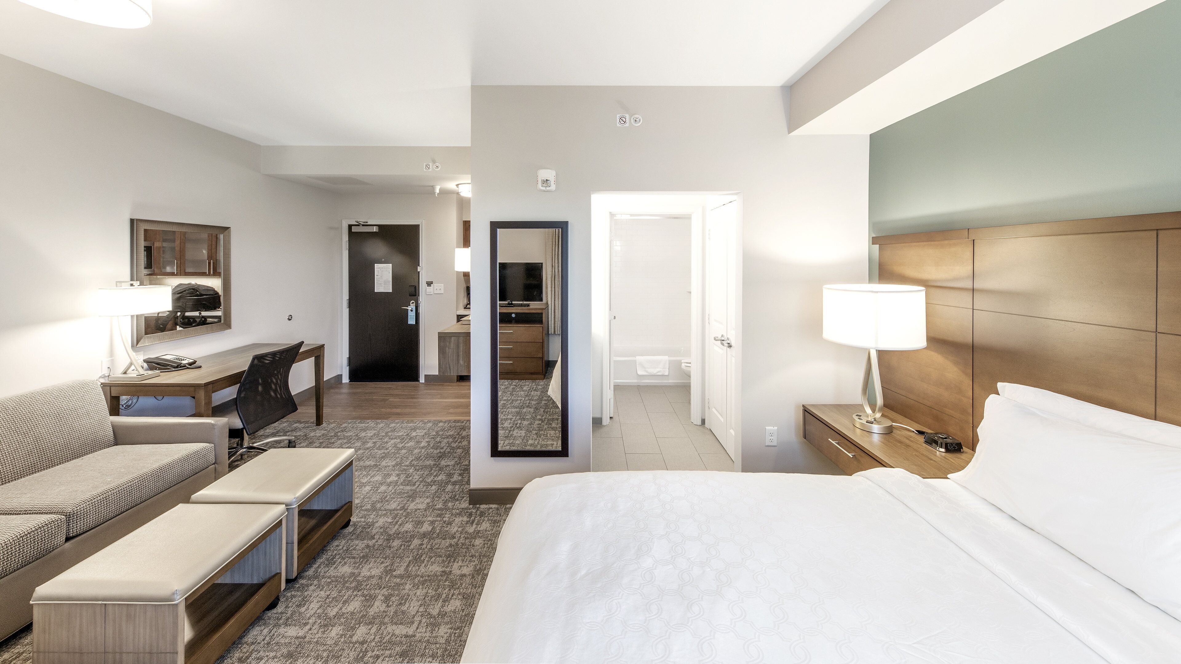 A hotel room with a bed, a desk, a chair, a sofa, a mirror, and a bathroom visible through an open door.
