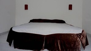 Uma cama de solteiro com lençol branco e cobertura marrom escura, posicionada contra uma parede branca lisa com duas pequenas arandelas vermelhas.
