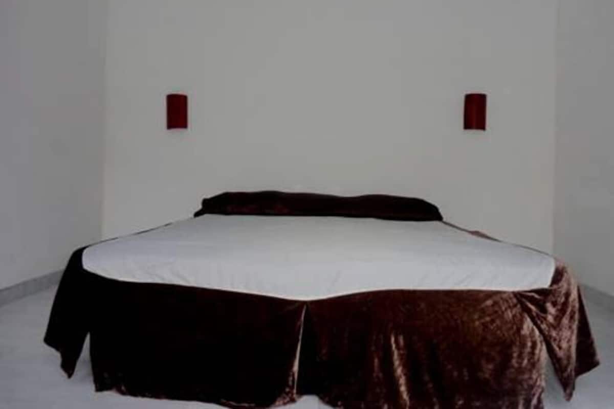 A single bed with a white sheet and a dark brown cover, positioned against a plain white wall with two small red wall sconces.