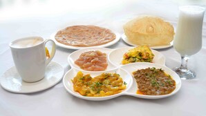 A breakfast setting with a cup of coffee, a plate of flatbread, a glass of milk, and various dishes including a vegetable curry and a potato dish. - Hotel Avalon Suites (Multan)