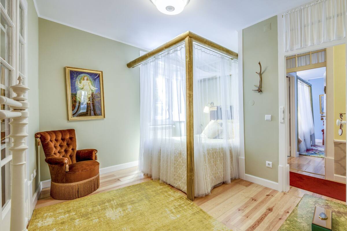 Une chambre avec un lit à baldaquin, un fauteuil capitonné, un tapis vert et un tableau au mur.