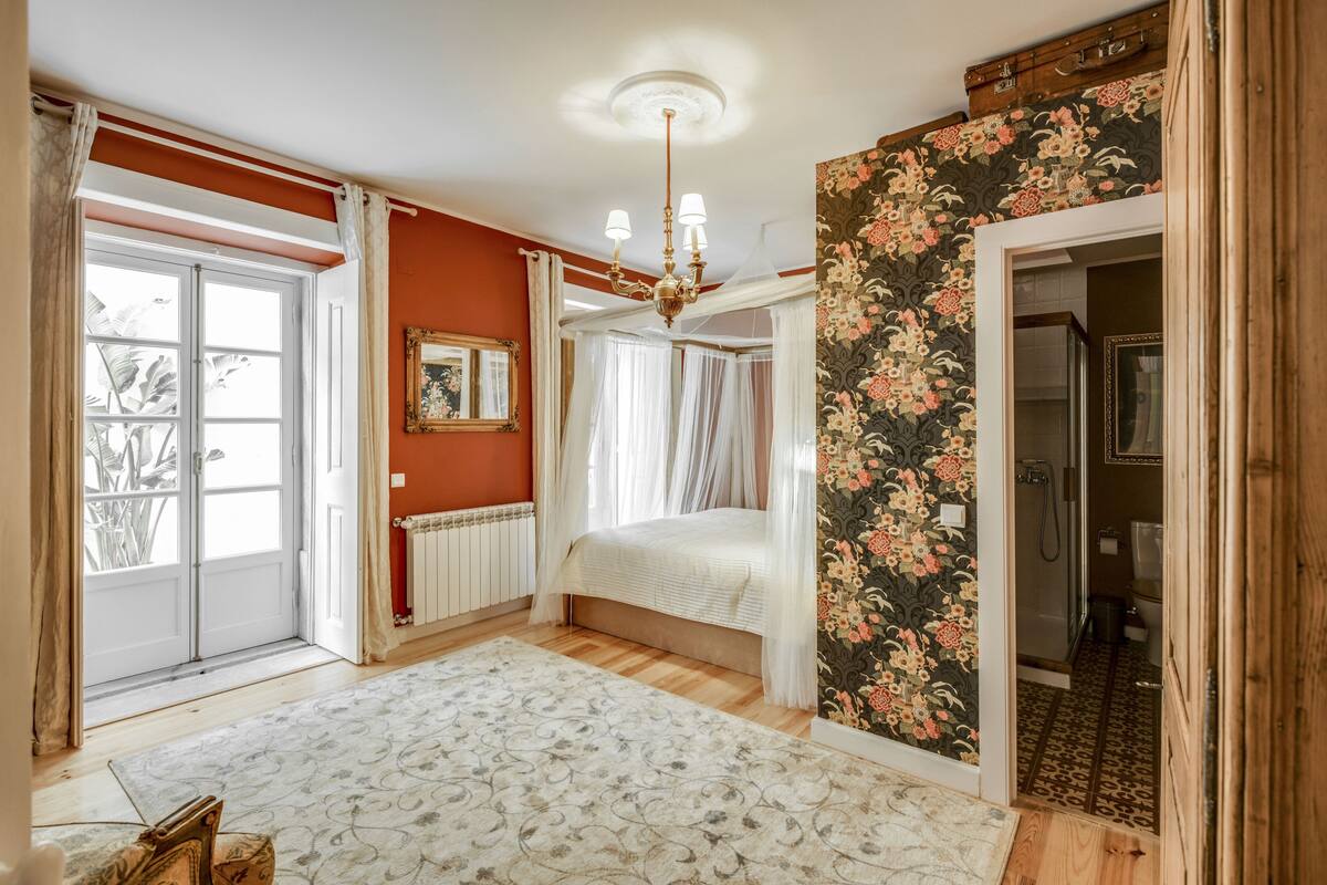 Une chambre à coucher avec un lit, un radiateur, une fenêtre avec des rideaux, un papier peint à motifs floraux et une porte en bois.