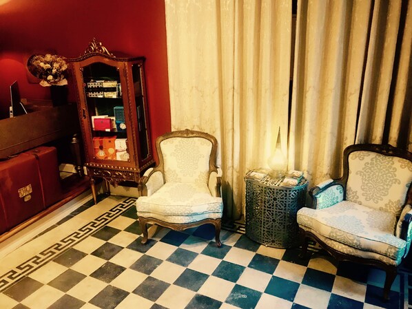 A room with a checkered floor, two armchairs, a side table with a lamp, a mirror, and a red cabinet.