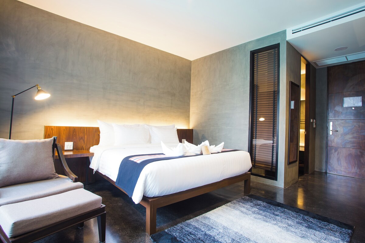 A modern hotel room with a large bed, a bedside table, a chair, and a lamp.