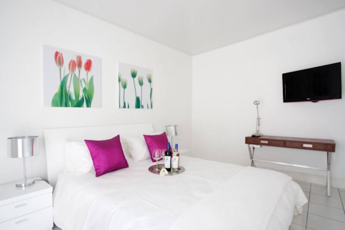 A modern hotel room with a large bed, white bedding, two purple pillows, a bedside table with a lamp, a tray with wine bottles, and a flat-screen TV mounted on the wall.
