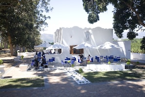 An outdoor seating area with white walls and blue chairs, surrounded by trees and potted plants.
