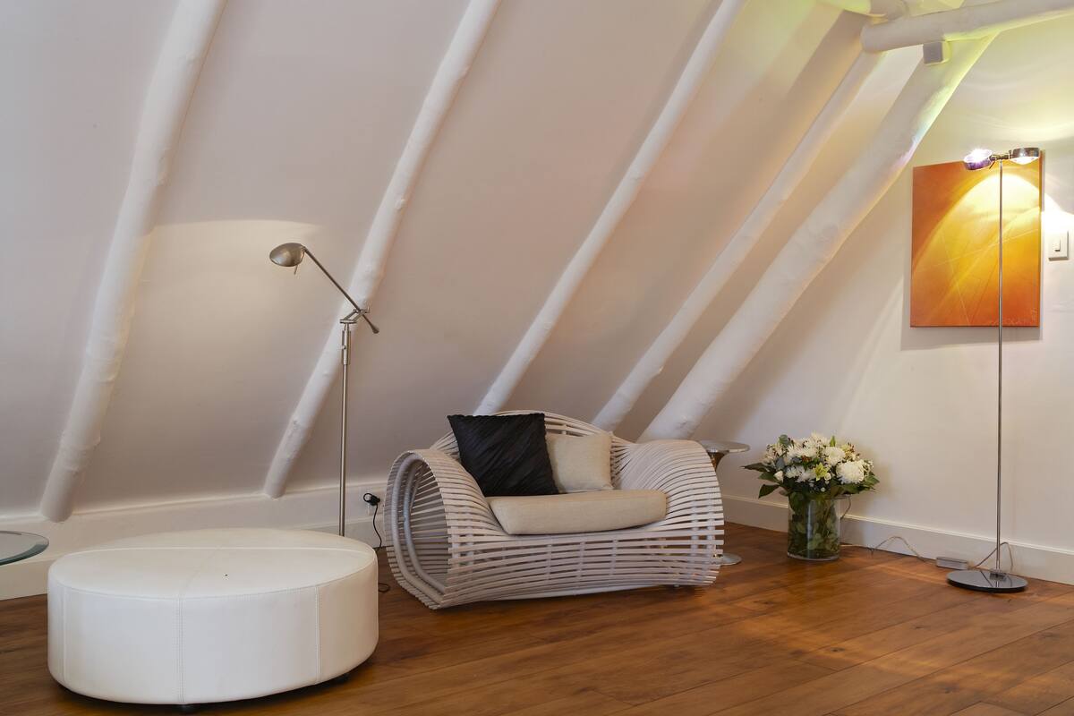 A cozy loft bedroom with a sloped ceiling, a wicker sofa, a round ottoman, a floor lamp, and a vase with flowers.