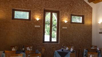 Un comedor con mesas y sillas de madera, paredes de piedra y ventanas con vistas a la vegetación.