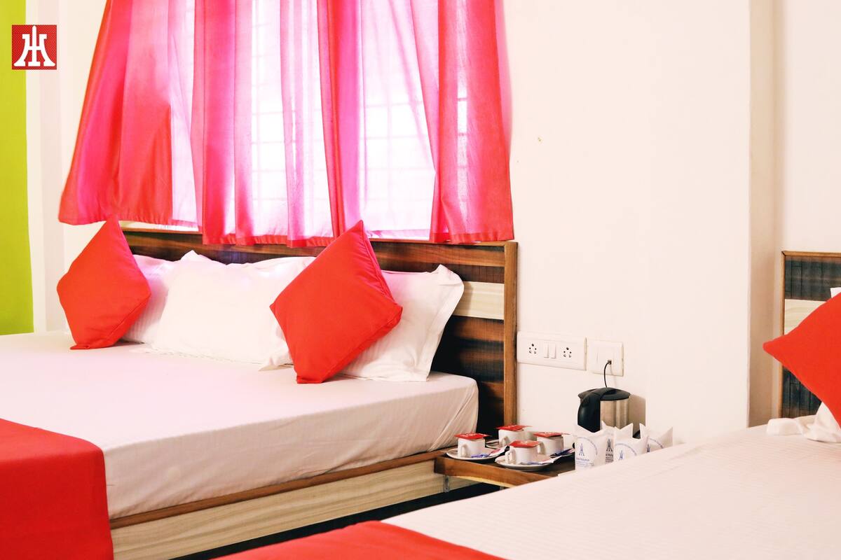 A hotel room with a bed, pillows, a bedside table with cups and a kettle, and a window with curtains.