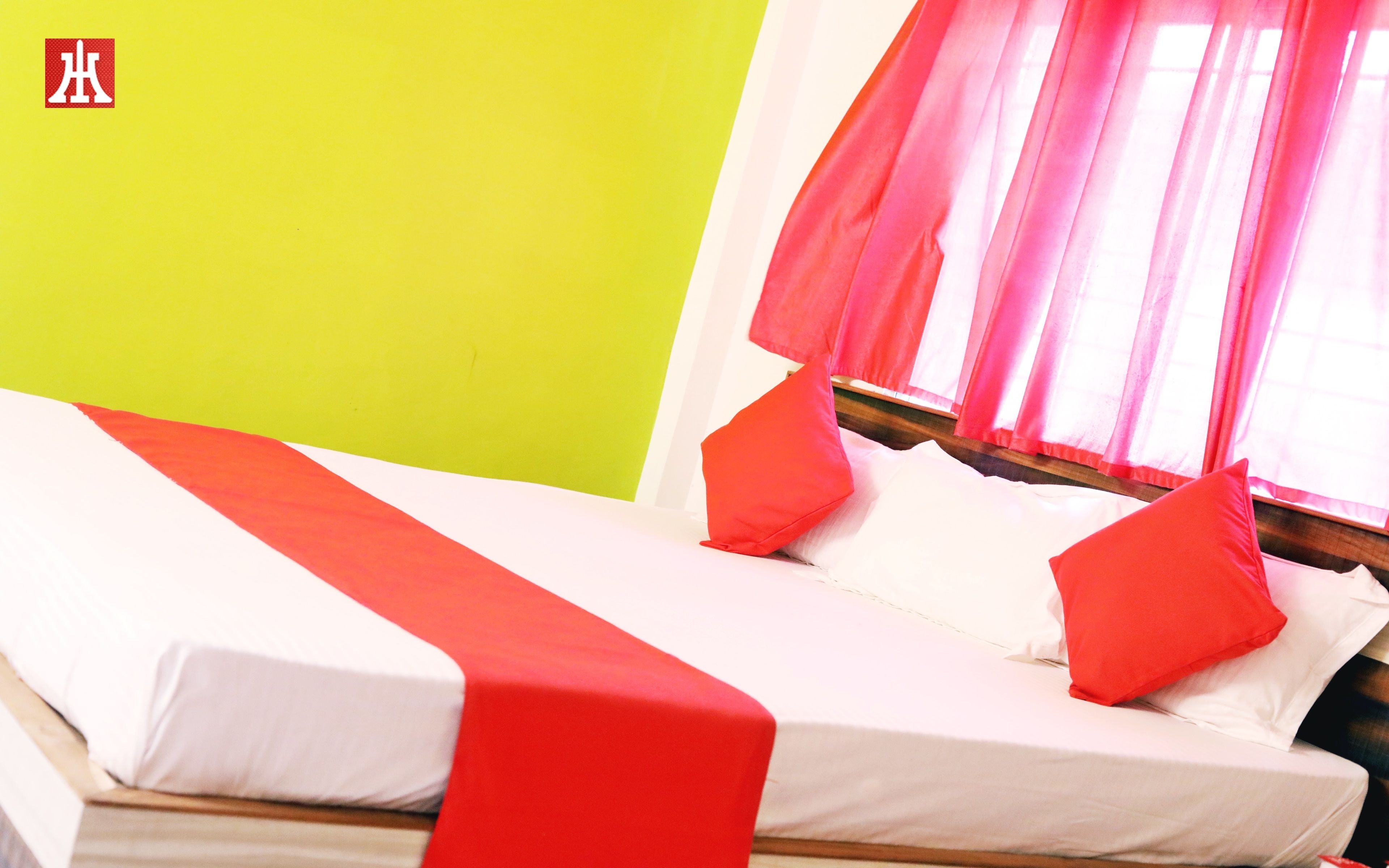 A bed with white bedding and red accents, a green wall, and a window with pink curtains.