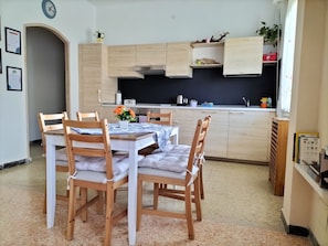 Un coin repas avec une table dressée pour quatre personnes, des chaises en bois, une cuisine en arrière-plan et une horloge au mur.
