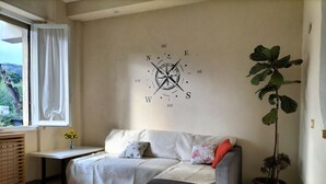 A cozy living room with a sofa, a small table with a vase of flowers, a large wall clock, and a potted plant.