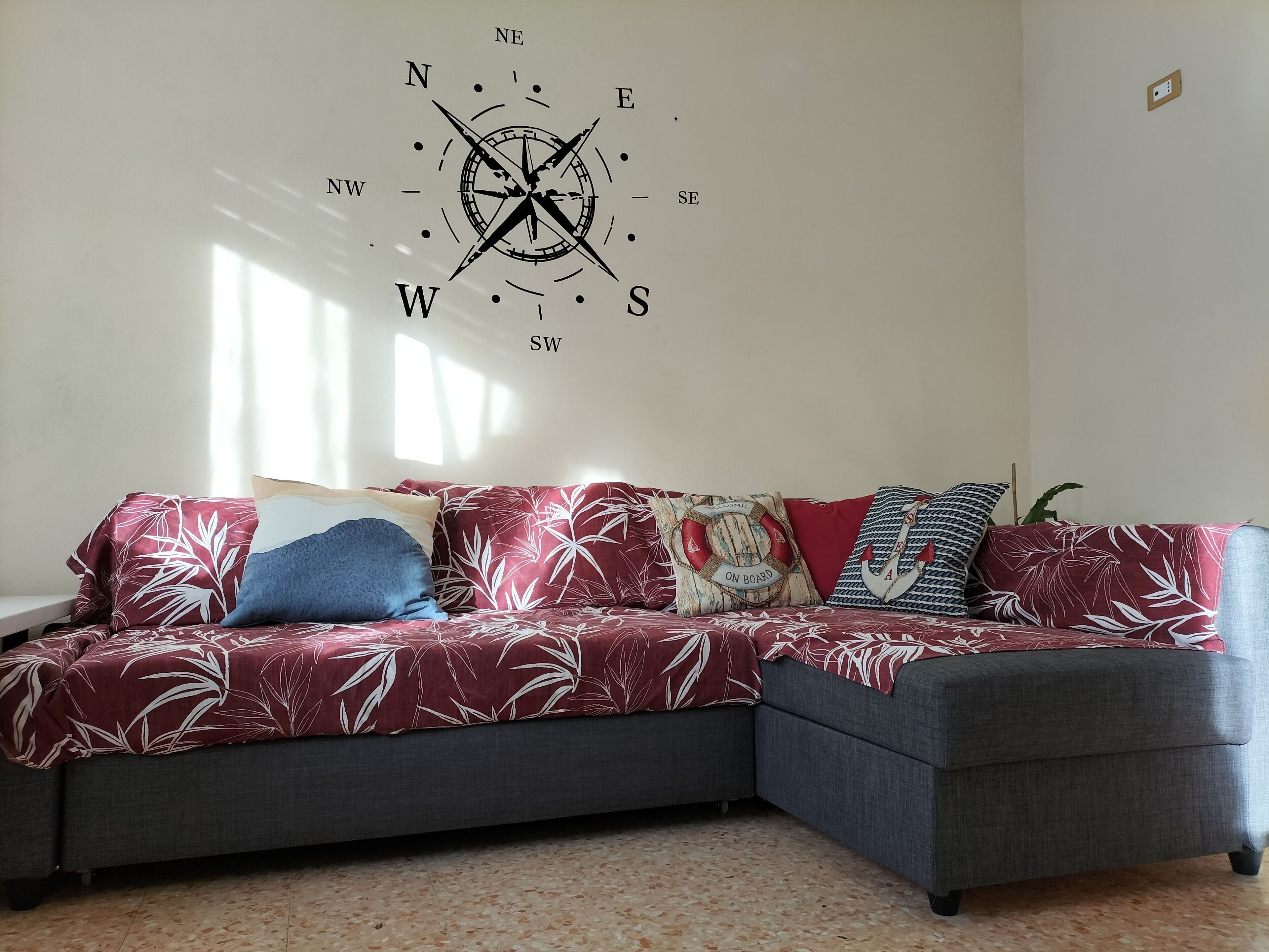 A corner sofa with a red and white leaf pattern, a compass wall decoration, and a small plant in the corner.
