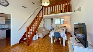 A spacious living area with a wooden staircase, a dining table set for four, and a kitchen visible in the background. - Cozy House Near Dunes Beach, 2 Bedrooms + Mezzanine, Parking & Garden, Baby Gear Included (Bretignolles-sur-Mer)