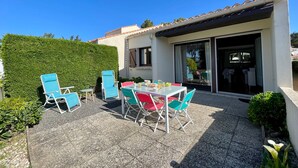 A patio with a table and chairs, a hedge, and a building with a tiled roof. - Cozy House Near Dunes Beach, 2 Bedrooms + Mezzanine, Parking & Garden, Baby Gear Included (Bretignolles-sur-Mer)